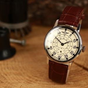 May include: A vintage-style wristwatch with a cream-colored face, black numerals, and a brown leather band. The watch features a small seconds dial and a silver-toned case. The watch is set against a wooden background.