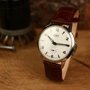 Könnte beinhalten: Eine Vintage-Armbanduhr mit weißem Zifferblatt, schwarzen Zeigern und goldfarbenen Ziffern. Die Uhr hat ein braunes Lederarmband und das Wort "СТАРТ" ist auf dem Zifferblatt aufgedruckt. Die Uhr liegt auf einer Holzoberfläche.