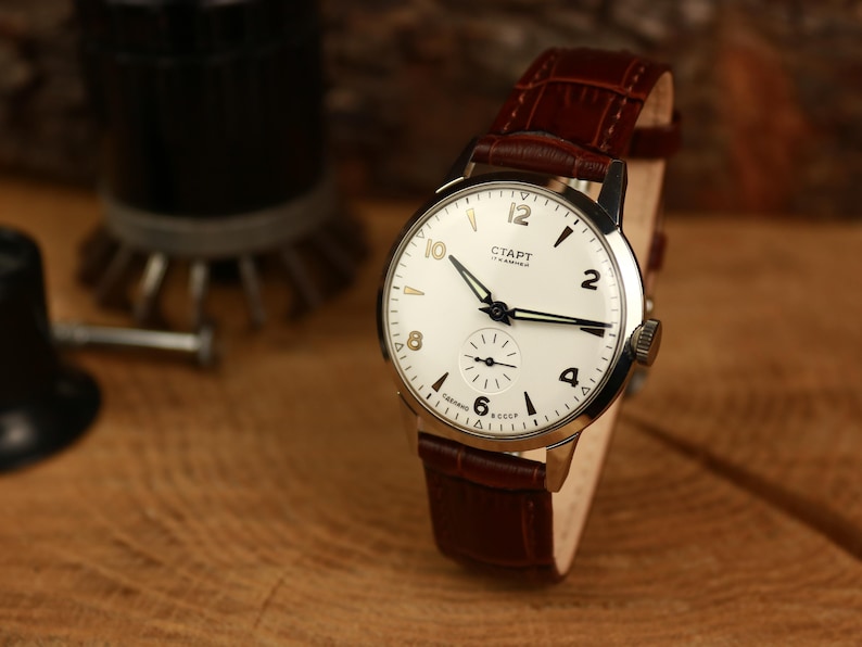 May include: A vintage wristwatch with a white face, black hands, and gold-toned numerals. The watch has a brown leather strap and the word "СТАРТ" is printed on the face. The watch is displayed on a wooden surface.