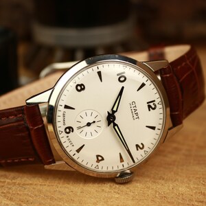 May include: A classic wristwatch featuring a cream dial, black hands, and a sub-dial for seconds. The watch has a stainless steel case and a brown leather strap. The watch face displays the text "CTAPT" and "17 KAMHEЙ".
