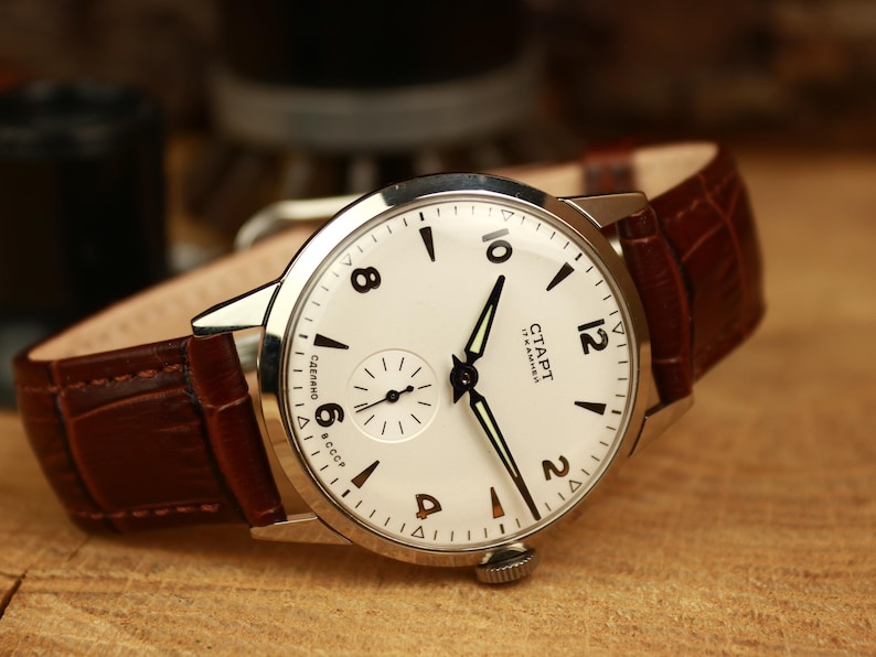 May include: A classic wristwatch featuring a cream dial, black hands, and a sub-dial for seconds. The watch has a stainless steel case and a brown leather strap. The watch face displays the text "CTAPT" and "17 KAMHEЙ".
