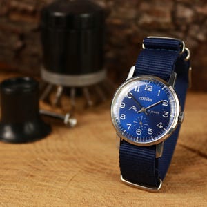 Puede incluir: Un reloj de pulsera vintage con esfera azul y correa de nailon azul marino. La esfera del reloj presenta números y manecillas blancas, con la marca "CORNAVIN". El reloj está sobre un fondo de madera, con herramientas de reparación de relojes en el fondo.