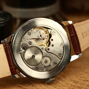 May include: Detailed view of a vintage wristwatch's inner workings. The silver watch face displays gears, springs, and jewels. It features a brown leather strap. The watch is set against a blurred background.