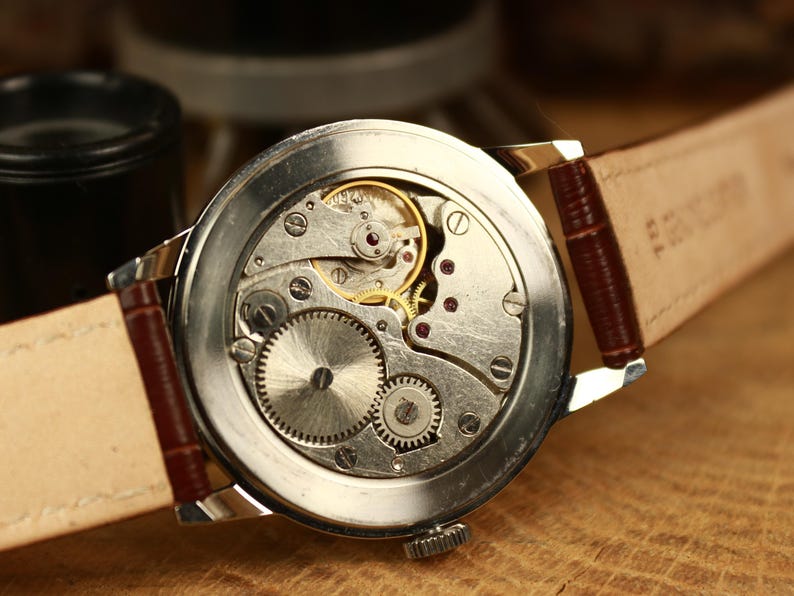 May include: Detailed view of a vintage wristwatch's inner workings. The silver watch face displays gears, springs, and jewels. It features a brown leather strap. The watch is set against a blurred background.