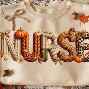 May include: Cream-colored sweatshirt with the word "NURSE" in a fall-themed design. The letters are textured with knitted patterns, pumpkins, acorns, and fall leaves. The letter "E" has a leopard print pattern.