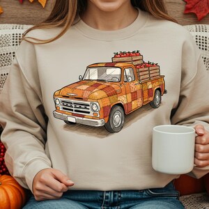 May include: A beige sweatshirt featuring an illustration of an orange vintage pickup truck with a patchwork design. The truck bed is filled with red apples. The image evokes a fall harvest theme.