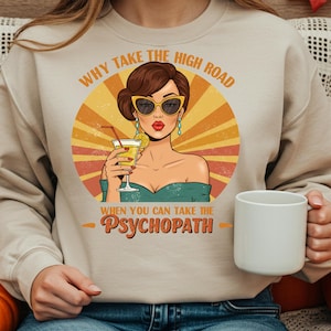 Op de afbeelding: Beige sweatshirt met een retro-illustratie van een vrouw met een zonnebril die een cocktail vasthoudt. De afbeelding bevat de tekst "WHY TAKE THE HIGH ROAD WHEN YOU CAN TAKE THE PSYCHOPATH."
