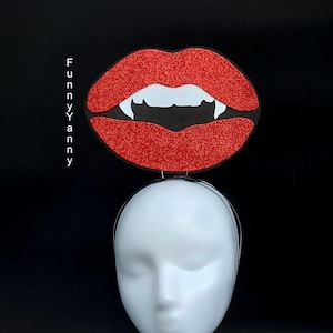 May include: A Halloween headband with a large, glittery red lip design and white fangs. The lips are mounted on a thin, silver headband. The text "FUNNYYANNY" is printed vertically on the left side of the image. The headband is displayed on a white mannequin head against a black background.