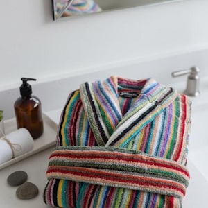 Könnte beinhalten: Ein gefalteter Bademantel aus Frottee mit bunten Streifen in Rot, Grün, Blau und Gelb. Der Bademantel liegt auf einer weißen Arbeitsplatte. Ein brauner Seifenspender und gerollte Handtücher sind ebenfalls zu sehen.