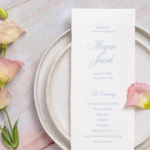 Op de afbeelding: Een trouwprogramma met de namen "Megan & Jacob" en ceremoniedetails, geplaatst op een wit bord. Roze en gele bloemen en een marmeren achtergrond maken de elegante scène compleet.