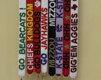 May include: A collection of beaded straps in various colors, each displaying team names like "GO BEARCATS!" and "CHIEFS KINGDOM." The straps hang from a metal bar, showcasing a variety of school spirit designs with gold-colored clasps.
