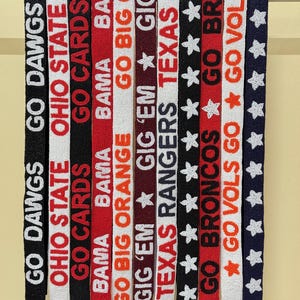 May include: A collection of beaded straps in various colors, each featuring team names and phrases like "GO DAWGS," "OHIO STATE," and "TEXAS RANGERS." The straps are adorned with white stars and gold-colored clasps.