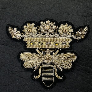 Puede incluir: Parche bordado con una abeja coronada. La abeja está detallada con hilo dorado y rayas negras. La corona también es dorada con detalles de gemas transparentes. Sobre un fondo negro.