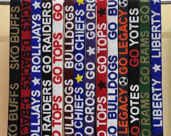 Könnte beinhalten: Eine Sammlung von Perlen-Lanyards mit Teamnamen und Sätzen wie "GO TOPS" und "GO RAIDERS". Die Lanyards sind in verschiedenen Farben, darunter Schwarz, Rot, Weiß und Blau, und hängen an einer Metallstange.