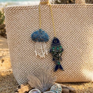 Pode incluir: Uma sacola tote bege com padrão de espinha de peixe, corrente dourada e pingentes de contas em forma de água-viva e peixe. A sacola está na areia com conchas, madeira flutuante e um leque do mar. O oceano é visível ao fundo.