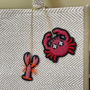 May include: A beige tote bag with a herringbone pattern, featuring two beaded charms: a pink lobster and a red crab with black accents. The charms are suspended by gold cords.