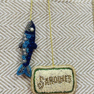 May include: Two beaded ornaments: a blue fish and a rectangular sardine can. The fish is blue with a pearl eye. The sardine can is off-white with the word "SARDINES" in gold beads, framed by green and gold beads.