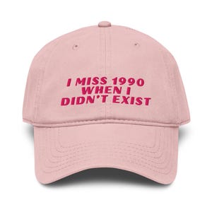 May include: A light pink baseball cap with the text "I MISS 1990 WHEN I DIDN'T EXIST" in a bold, pink font. The cap has a curved brim and a button on top.