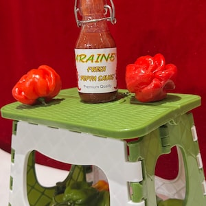 May include: A small bottle of Arraine Fresh Peppa Sauce with a white label and a red liquid, sits on a green and white folding stool. Two red peppers are on the stool. The background is red.