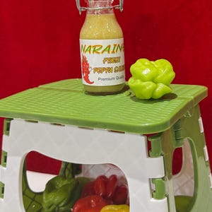 May include: A small glass bottle of "NARAIN FRESH PEPPA SAUCE" with a red chili pepper logo, sits on a green and white folding step stool. A green pepper is next to the bottle. The stool contains various colorful peppers.