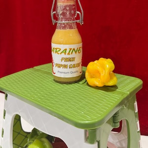 May include: A small glass bottle of "Araine Fresh Peppa Sauce" with a white label and a metal clasp closure. The bottle sits on a green and white folding stool next to a yellow pepper. The background is a solid red.