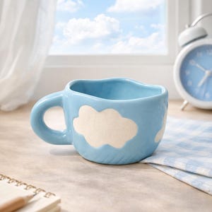 Könnte beinhalten: Hellblaue Keramiktasse mit Wolkenmuster. Die Tasse hat einen Henkel und einen wellenförmigen oberen Rand. Ein weißer Wecker und ein Notizbuch befinden sich im Hintergrund.