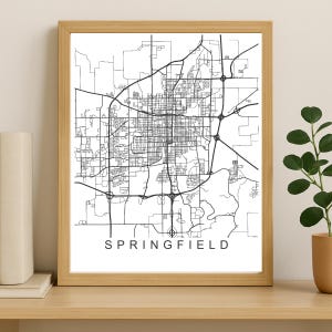 Puede incluir: Una impresión enmarcada en blanco y negro del mapa de Springfield, con una red de carreteras detallada. El nombre de la ciudad está impreso en una fuente limpia y sans-serif en la parte inferior. La impresión está en un marco de madera marrón claro.