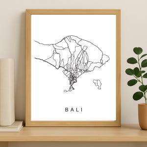 Może przedstawiać: Oprawiony wydruk z minimalistyczną czarno-białą mapą Bali. Mapa przedstawia zarys wyspy i sieć dróg. Słowo "BALI" jest wydrukowane poniżej. Rama jest w jasnym kolorze drewna.