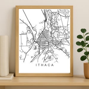 May include: A framed black and white map print of Ithaca, New York. The map features a detailed outline of the city's streets and layout, with the city name printed in a clean, sans-serif font below the map. The print is in a light brown wooden frame.