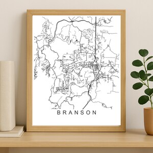 May include: A framed black and white map print of Branson, Missouri. The map details the city's roads and infrastructure. The word "BRANSON" is printed in a simple, sans-serif font at the bottom of the print. The print is in a light-colored wooden frame.