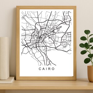 May include: A framed black and white map of Cairo, Egypt. The city's road network is depicted in detail against a white background. The word "CAIRO" is printed in black at the bottom of the image. The frame is made of light-colored wood.