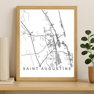 May include: A framed print showing a black and white map of Saint Augustine, Florida. The map depicts the city's roads and buildings. The text "SAINT AUGUSTINE" is printed at the bottom. The frame is light brown. The print is in a wooden frame.