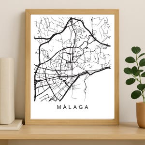 May include: A framed black and white map of Málaga, Spain. The city map is a detailed line drawing, set against a white background. The word "MÁLAGA" is printed in a bold, sans-serif font at the bottom of the print. The frame is a light wood color.