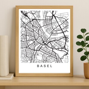 Peut inclure: Une impression encadrée en noir et blanc d'une carte de Bâle, en Suisse. La carte présente un plan détaillé des rues, avec le nom de la ville imprimé en bas dans une police sans-serif. L'impression est encadrée dans un cadre en bois naturel.