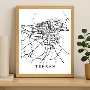 May include: A framed print featuring a black and white map of Tehran. The city's road network is depicted in detail. The word "TEHRAN" is printed in a clean, sans-serif font at the bottom of the print. The frame is a light brown color.