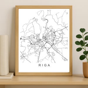 Op de afbeelding: Een ingelijste print met een zwart-wit kaart van Riga, Letland. De stadskaart staat op een witte achtergrond, met de naam van de stad in het zwart onderaan. De lijst is gemaakt van lichtgekleurd hout.