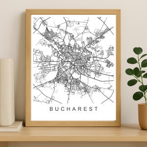 May include: A framed black and white map of Bucharest, Romania. The city map is detailed with roads and buildings. The word "BUCHAREST" is printed in a simple, sans-serif font at the bottom of the map. The frame is made of light-coloured wood.
