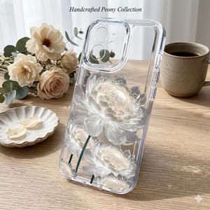 Könnte beinhalten: Transparente Handyhülle mit einem weißen Pfingstrosen-Design. Die Hülle hat einen transparenten Rand und den Text "Handcrafted Peony Collection". Das florale Design umfasst weiße Blütenblätter und grüne Stiele. Die Hülle liegt auf einer Holzoberfläche.