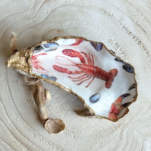 Op de afbeelding: Decoratieve oesterschelp met een goudkleurige rand en een kreeftontwerp. De schelp is wit met een rode kreeftillustratie en blauwe accenten. Een stuk drijfhout is aan de schelp bevestigd.