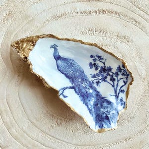 Op de afbeelding: Een decoratieve oesterschaal met een blauw pauwenontwerp. De binnenkant is wit met een blauwe pauw- en boomillustratie. De rand van de schelp is afgewerkt met goudverf. De schaal ligt op een lichtgekleurd oppervlak.