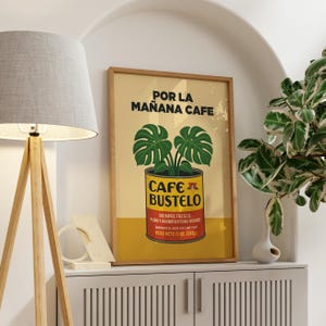 May include: A framed poster with a vintage design. The poster features the text "POR LA MAÑANA CAFE" and "CAFE BUSTELO" with a graphic of a plant in a can. The poster also includes the text "PESO NETO 10 OZ. (283g)".