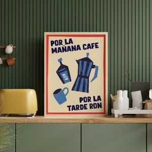 May include: A framed poster with a cream background features blue illustrations of a coffee pot, a bottle of rum, and a mug. The text reads "POR LA MAÑANA CAFE" and "POR LA TARDE RON" in blue.