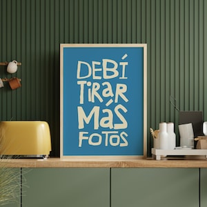 Op de afbeelding: Een ingelijste kunstdruk met een blauwe achtergrond en de Spaanse tekst "DEBÍ TIRAR MÁS FOTOS" in een vet, gebroken witte lettertype. De print wordt tentoongesteld op een houten plank met een gele broodrooster en decoratieve items tegen een groene muur.