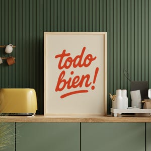 Op de afbeelding: Een ingelijste print met de woorden "todo bien!" in rood script op een crèmekleurige achtergrond. De print zit in een licht houten frame en staat op een houten plank met een gele broodrooster en witte keramische flessen.