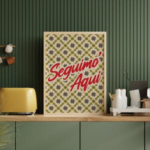 May include: A framed print with the words "Seguimo' Aqui" in red script. The background of the print features a repeating pattern of yellow, green, and brown star-like shapes. The print is displayed on a wooden shelf with a yellow toaster.