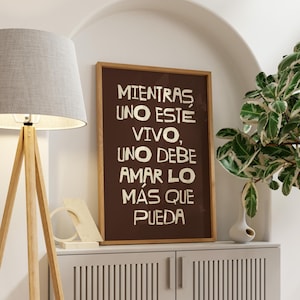 K&ouml;nnte beinhalten: Ein gerahmter Druck mit dem spanischen Text "Mientras uno est&eacute; vivo, uno debe amar lo m&aacute;s que pueda" in wei&szlig;en Buchstaben auf braunem Hintergrund. Der Rahmen ist holzfarben. Der Druck wird auf einem wei&szlig;en Schrank mit Lampe und Pflanze angezeigt.