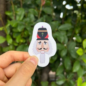 Puede incluir: Una pegatina con una ilustración de dibujos animados de un soldado cascanueces. El cascanueces tiene barba y bigote blancos, un sombrero rojo y negro y una expresión seria. La pegatina se sostiene en una mano sobre un fondo de hojas verdes.