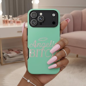 May include: A mint green phone case with a black camera module. The case features the text "Angel Bitch" in pink rhinestones, with a halo above the word "Angel". The case is held by a person with pink nails and a silver bracelet.