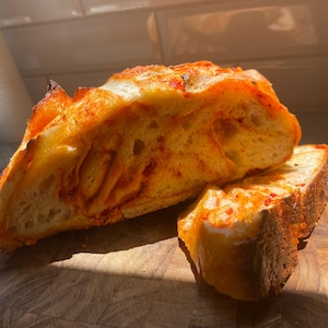 Puede incluir: Primer plano de una rebanada de pan, posiblemente focaccia, con un tono naranja vibrante, cortada para revelar la textura interior. El pan está colocado sobre una tabla de cortar de madera, con copos de pimiento rojo visibles.
