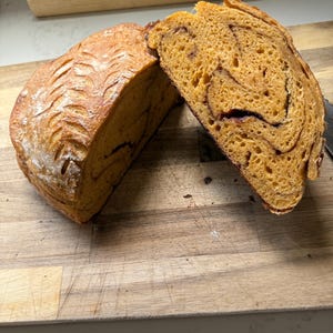 May include: A loaf of orange-tinted bread cut into two pieces, featuring a dark brown swirl pattern throughout the crumb. The crust has a golden-brown color with a decorative leaf-like pattern scored into the top surface of the bread.